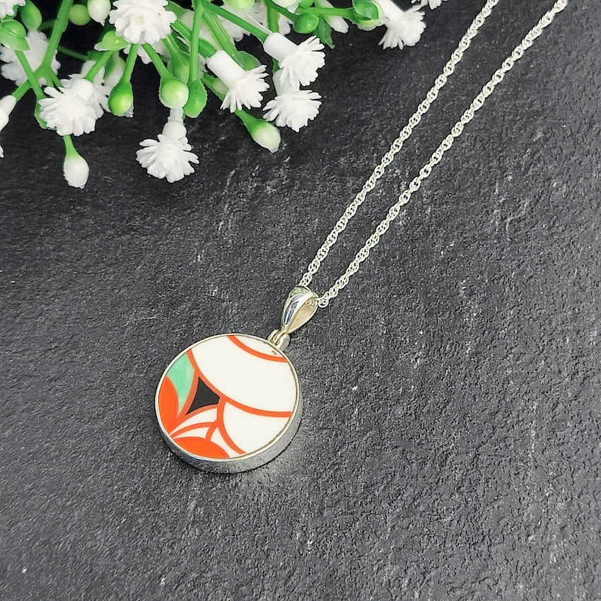 Clarice Cliff round pendant, 20mm, with orange, green, white and black. On a dark grey slate background.