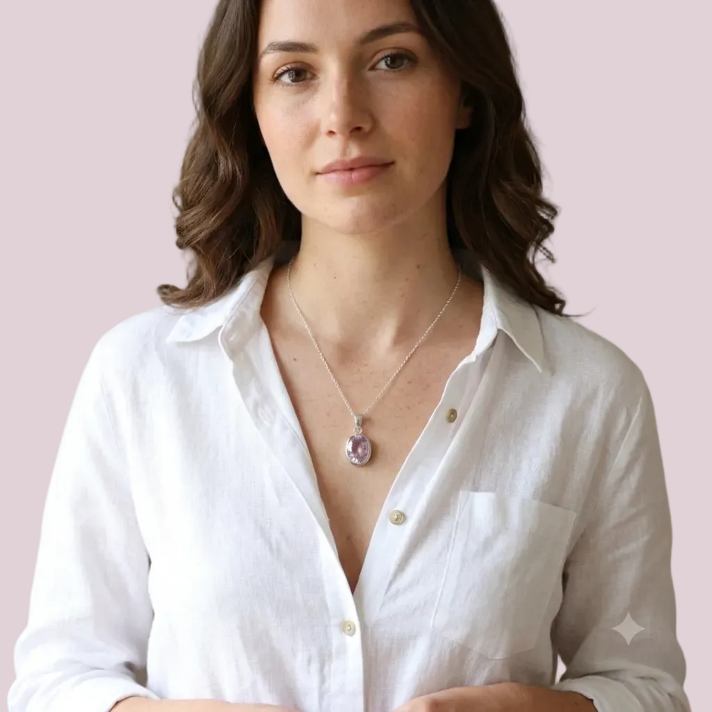 A woman wearing a silver-toned chain necklace with a lavender-purple oval pendant, paired with a white linen button-up shirt, set against a neutral beige background.