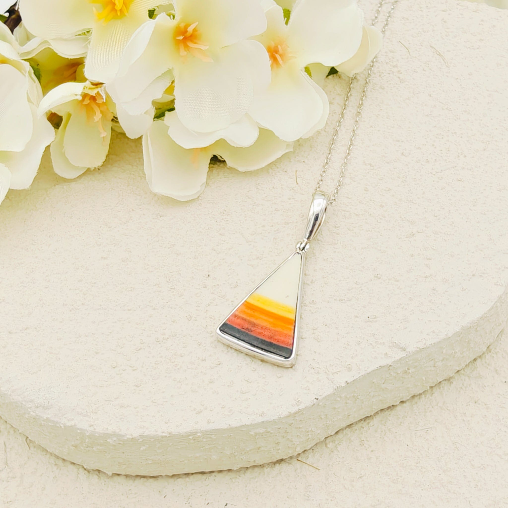 Clarice Cliff triangle pendant with a black base, yellow and orange stripes and white top point. It has a silver chain on a cream stone background with flowers above
