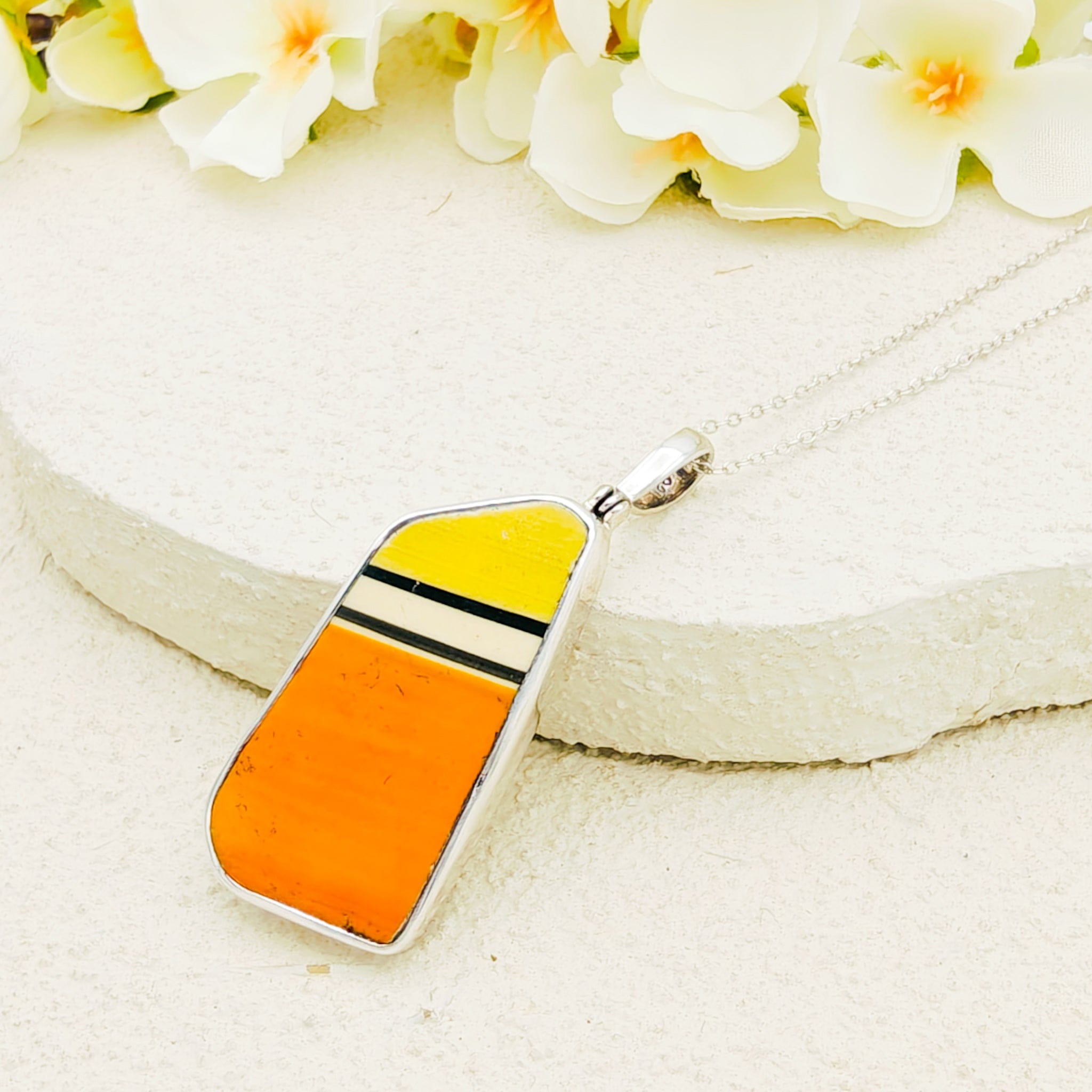 Colourful Clarice Cliff pendant with orange, white, black and yellow stripes, from the Fantasque collection, on a white surface with flowers in the background