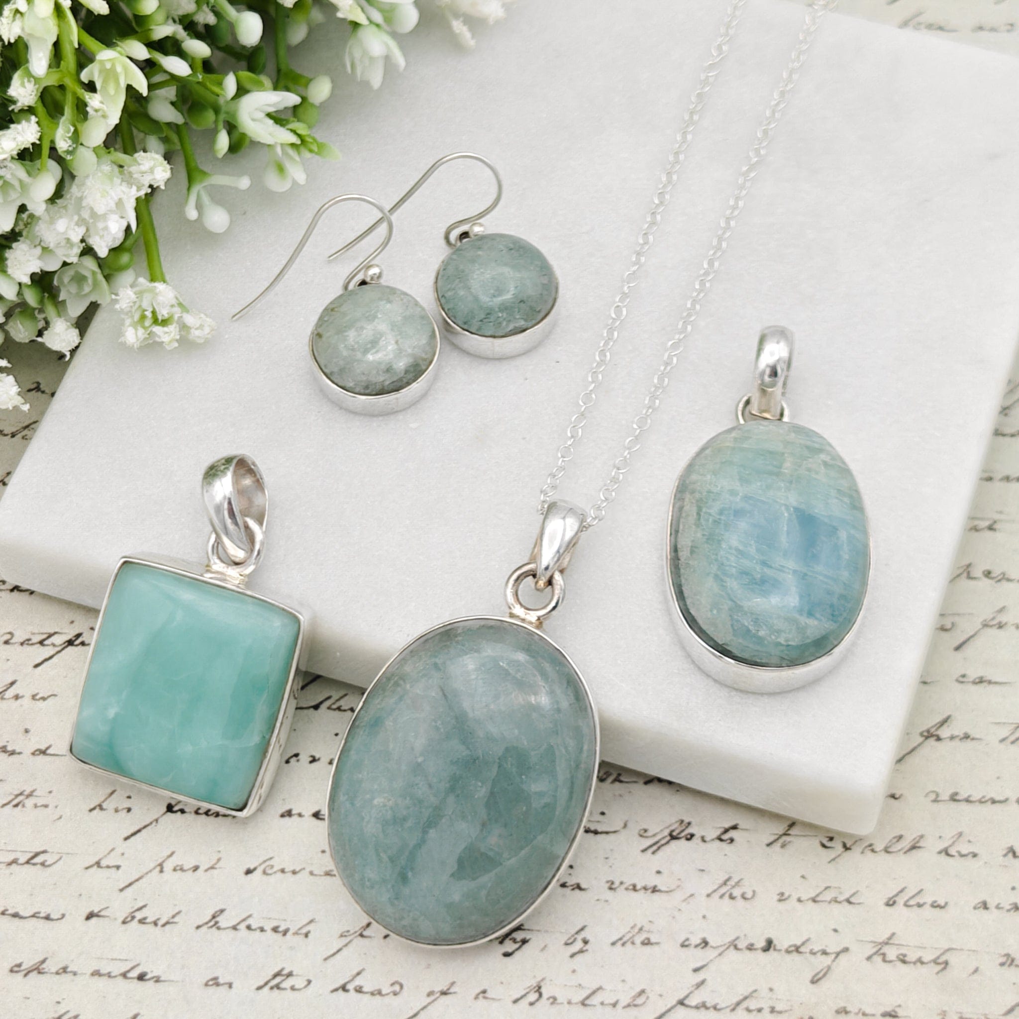 Selection of aquamarine pendants on a pale background of paper with faint writing on, and flowers in the background