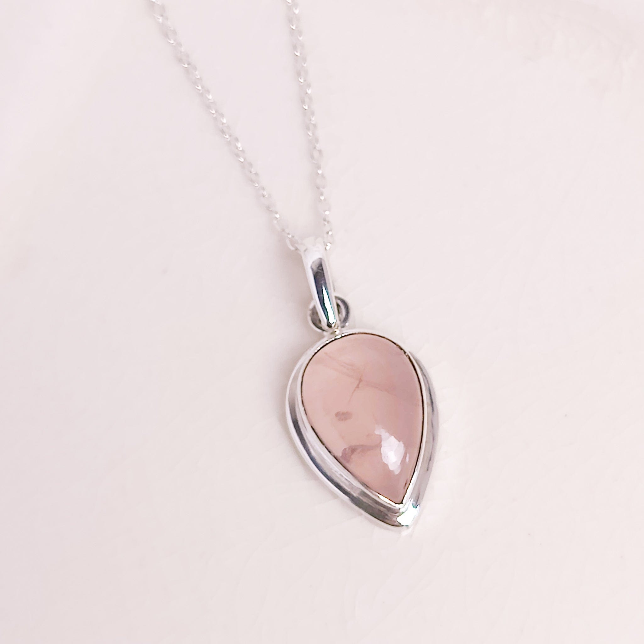 Rose quartz inverted teardrop with open back, allowing light to travel through the stone. It's on a pale pink background and has a silver chain through the bail