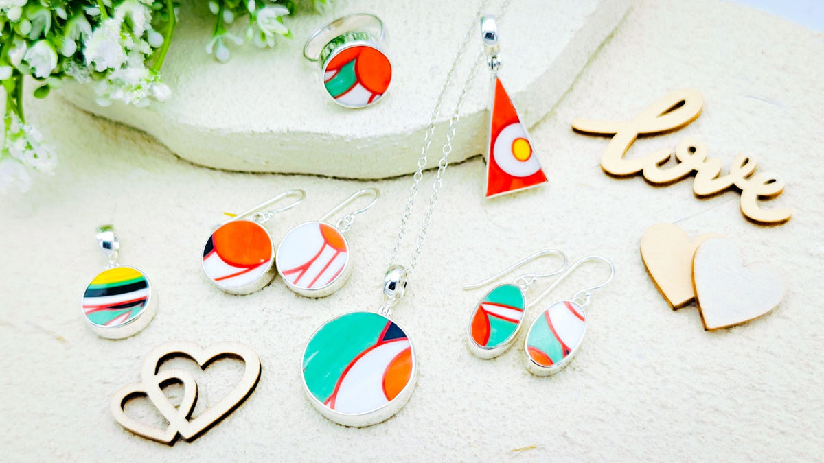 A styled flat-lay of colorful jewelry featuring round and teardrop pendants and matching earrings with abstract patterns in red, orange, teal, white, and yellow on a light textured background, alongside wooden heart cutouts and a 'love' sign.