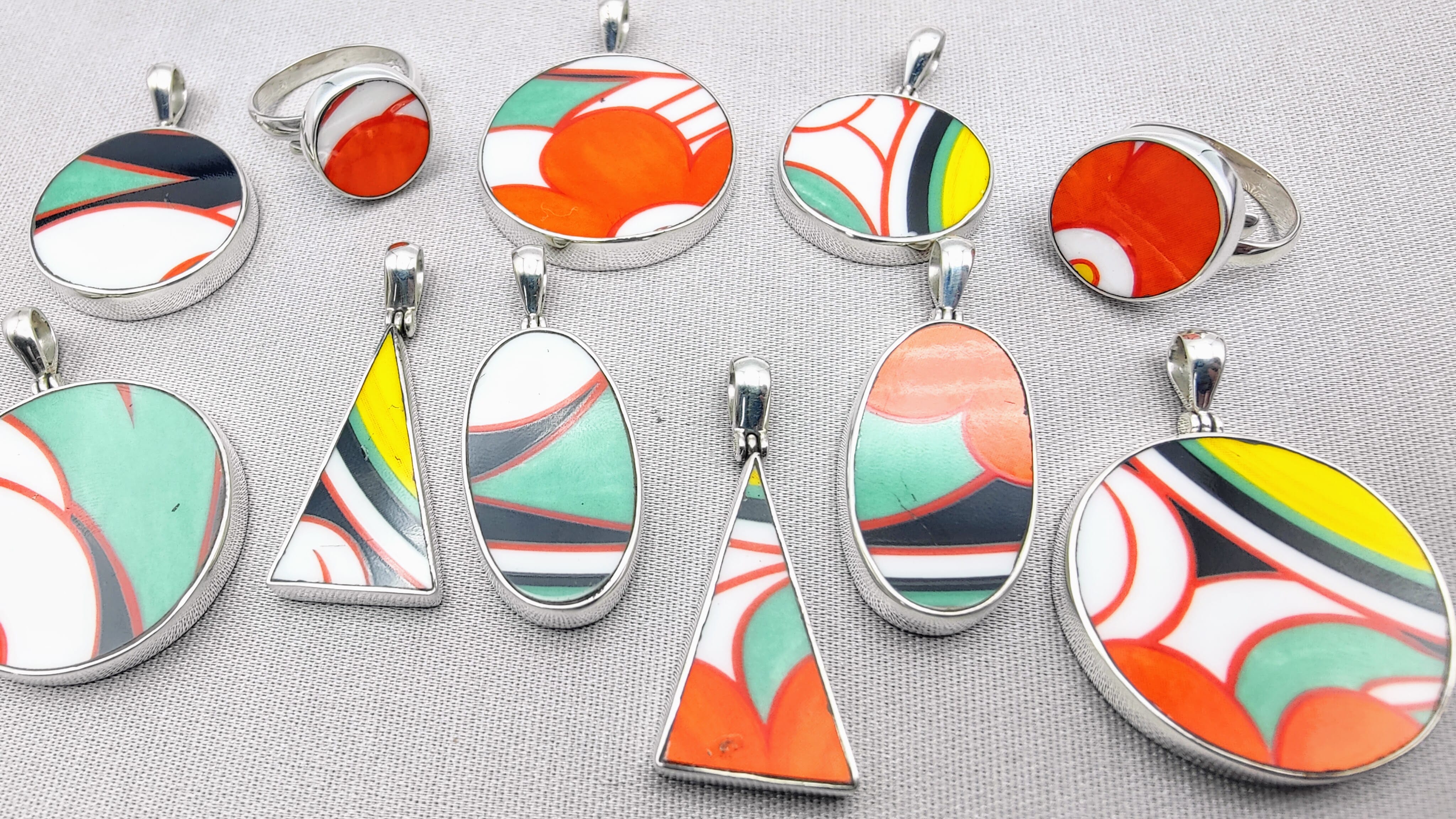 A set of colourful abstract-patterned Clarice Cliff jewellery pieces, including oval pendants and a ring, displayed on a light grey textured fabric background.