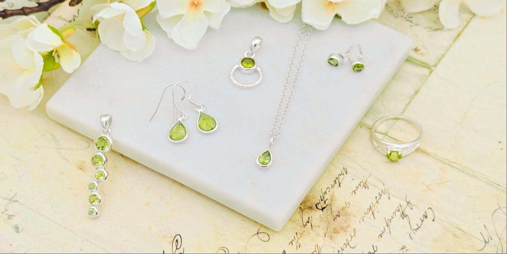 Silver jewellery set with green peridot gemstones, including a necklace, earrings, and rings, displayed on a white marble block with white flowers and vintage-style paper.