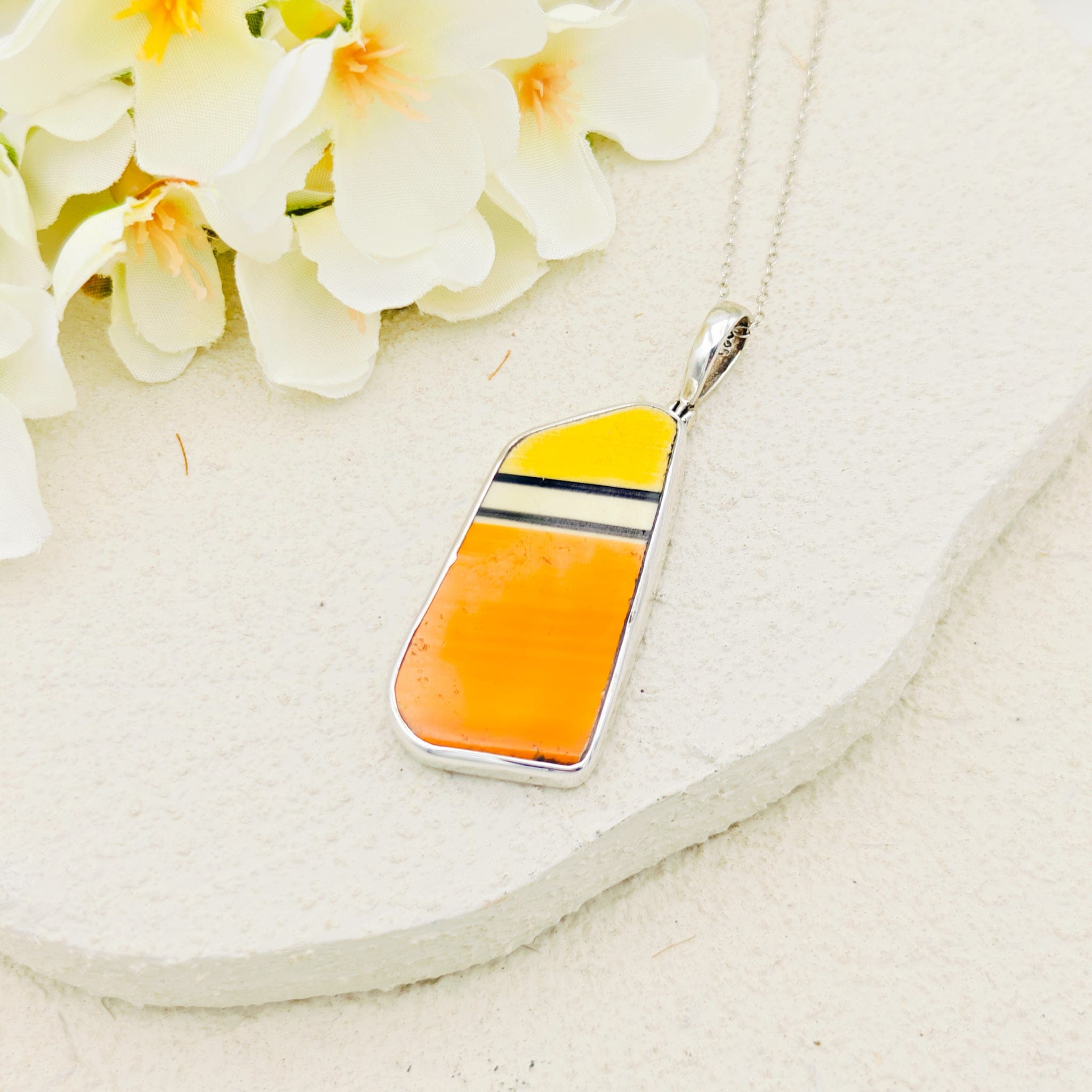 Orange and yellow Clarice Cliff pendant on a white background with flowers above. The pendant has a silver chain and black and white stripes on the ceramic fitting. 